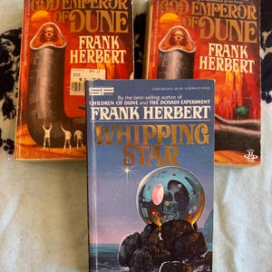 May include: Three science fiction books by Frank Herbert are displayed. Two copies of "God Emperor of Dune" are visible, featuring a red and orange cover with a central figure. The third book, "Whipping Star," has a blue cover with a space-themed illustration.