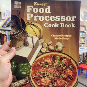 Puede incluir: Un libro de cocina vintage titulado "Sunset Food Processor Cook Book" con el subtítulo "Classic Recipes Made Easy". La portada presenta una pizza con ingredientes, un procesador de alimentos, verduras y una calabaza amarilla. El libro es sostenido por una persona.