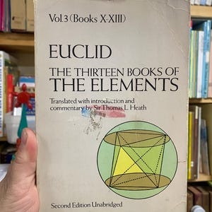 以下が含まれることがあります： 「ユークリッド原論」第3巻、第二版無修正版の使い古されたコピー。表紙には、緑色の円の中に幾何学的な図が描かれています。本は手で持たれており、背景には本棚があります。