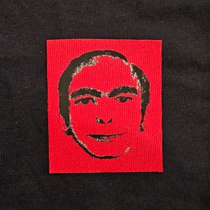 May include: A rectangular fabric patch featuring a black screen-printed graphic of a person's face on a vibrant red background. The patch is sewn onto a dark black textile surface, displaying a high-contrast portrait with distinct facial features.