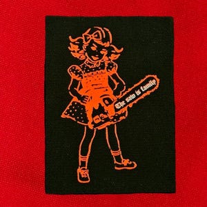 May include: A black and orange patch featuring a cartoon girl holding a chainsaw. The text "The saw is family" is written on the chainsaw.