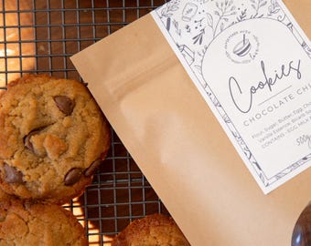 Może przedstawiać: Brązowa papierowa torba z białą etykietą, na której widnieje napis "Cookies Chocolate Chip" i lista składników. Etykieta ma kwiatowy wzór i małą ilustrację miski z łyżką. Torba ma przezroczyste okienko, przez które widać dwa ciasteczka z kawałkami czekolady.