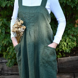 May include: A dark olive green pinafore apron dress featuring wide shoulder straps, a square neckline, and two large patch pockets on the front. The garment has vertical pleats below the chest and is worn over a long-sleeved white shirt.