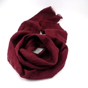 May include: A burgundy-colored scarf made of soft fabric. The scarf is draped in a circular pattern, with a fringed edge visible at the top. The scarf is on a white background.