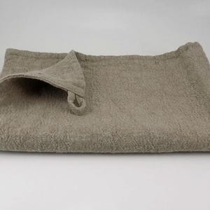 May include: A beige linen tea towel with a loop handle. The towel is folded in half and has a textured surface.