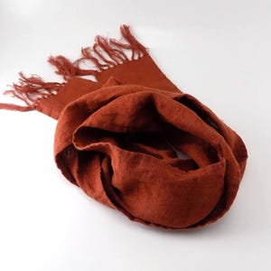 May include: A rust-colored linen scarf with fringe on the ends. The scarf is folded and arranged on a white background.