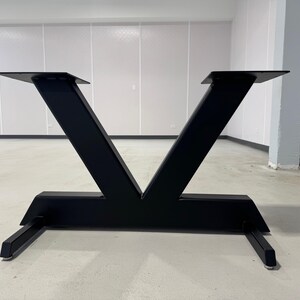 May include: Black metal table base with a V-shaped design. The base has a rectangular platform and two legs that extend out from the platform.