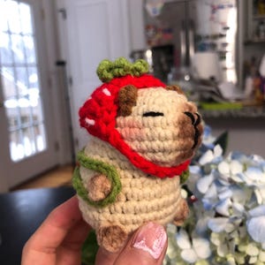 May include: A crocheted stuffed toy wearing a red and green strawberry hat. The toy is cream coloured with brown eyes and a brown nose.