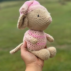 Op de afbeelding: Een handgemaakte gehaakte lammetjespop in beige en roze. Het lam heeft een roze en beige gestreept lichaam, een bijpassende hoed en kleine zwarte ogen. De pop wordt in een hand gehouden tegen een vage groene achtergrond.