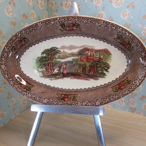 May include: An oval serving dish with a brown and white floral pattern and a scene of a castle and a river. The dish is on a silver stand.