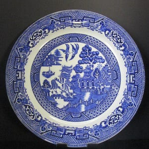 May include: A round, white and blue ceramic plate with a Willow pattern design. The plate features a detailed landscape scene with bridges, buildings, and birds. The border has a geometric and floral pattern. A classic piece of tableware.
