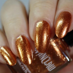 May include: Close-up of polished fingernails with a shimmering, metallic copper-gold finish. The nail polish has a glittery texture, reflecting light. A bottle of nail polish is partially visible in the background.