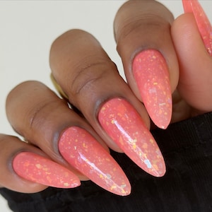 May include: Close-up of a hand with long, almond-shaped nails. The nails are painted a coral color with iridescent glitter flakes. The nails are a glossy finish and appear to be professionally done.