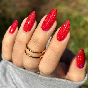 May include: Close-up of a hand with long, red glitter nail polish. The nails are almond-shaped and feature a vibrant, sparkling finish. A gold double-band ring adorns the ring finger. The background is blurred, with hints of green.