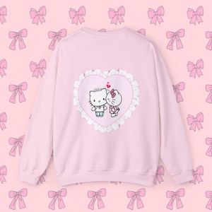 Puede incluir: Sudadera rosa con un diseño de corazón blanco con Hello Kitty y Dear Daniel besándose. El corazón tiene un borde de encaje blanco.