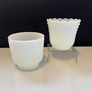 Könnte beinhalten: Zwei weiße Glasvasen. Eine Vase hat ein Spiraldesign, die andere ein strukturiertes, gepunktetes Muster. Beide sind undurchsichtig und stehen auf einer weißen Oberfläche mit schwarzem Hintergrund. Dekorative Vasen.