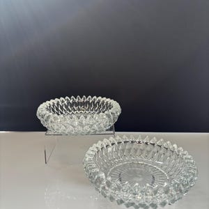 Könnte beinhalten: Zwei Aschenbecher aus klarem Glas mit Rautenmuster. Ein Aschenbecher steht auf einem transparenten Ständer, der andere auf einer weißen Oberfläche. Die Aschenbecher haben eine runde Form mit einem strukturierten Rand.