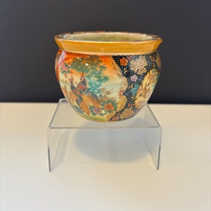 Peut inclure: Un bol décoratif en céramique avec un motif de paon. Le bol présente un motif floral et aviaire coloré avec des teintes orange, vertes et bleues. Le bord est de couleur jaune chaud. Le bol est présenté sur un présentoir en acrylique transparent.