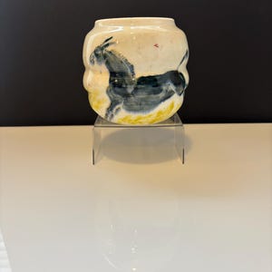 Puede incluir: Un jarrón de cerámica de color crema con forma redondeada. El jarrón presenta una imagen pintada de un burro azul oscuro con detalles amarillos en la base. El jarrón se exhibe sobre un soporte acrílico transparente.