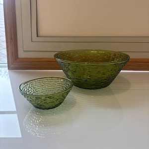 May include: Two green glass bowls with a textured, woven-like pattern. The bowls are different sizes, with the larger one in the background and the smaller one in the foreground. The bowls are sitting on a white surface.