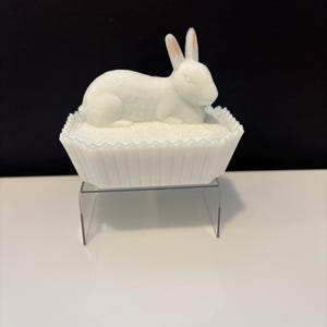 Puede incluir: Una figura de conejo de cerámica blanca con orejas rosadas descansa sobre un plato blanco con borde festoneado. El conejo está acostado y el plato está sobre un soporte acrílico transparente. El fondo es blanco y negro.