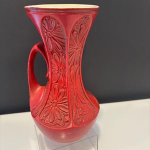 Puede incluir: Un jarrón de cerámica rojo con asa y boca ancha. El jarrón presenta un motivo floral tallado en la superficie. El interior es de color crema. El jarrón se exhibe sobre un soporte acrílico transparente.