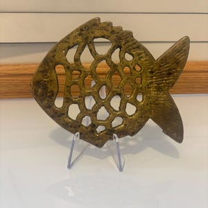 May include: A golden-coloured, openwork fish sculpture. The fish features a detailed, textured surface with circular cutouts. It is displayed on a clear acrylic stand, highlighting its intricate design and craftsmanship.