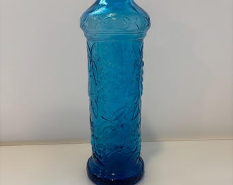 May include: A tall, cylindrical glass bottle in a vibrant shade of blue. The bottle features a narrow neck and a flared rim. The body is decorated with an embossed pattern of grapes and leaves, with a band of small floral motifs near the shoulder.