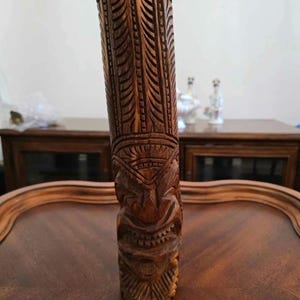 Puede incluir: Una estatua tiki de madera marrón oscuro con tallas intrincadas. La estatua tiene una cara humanoide con una boca ancha y ojos grandes.