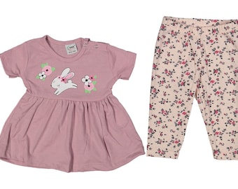 Puede incluir: Un conjunto de dos piezas para bebé con un vestido rosa de manga corta con un conejito blanco con una corona de flores y detalles florales. El conjunto también incluye un pantalón de color crema con estampado floral.