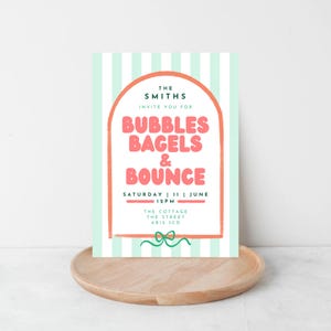 May include: A party invitation with a mint green and white striped background. The invitation features a coral-colored arch with the text "BUBBLES BAGELS & BOUNCE" in pink. The invitation is on a wooden tray.