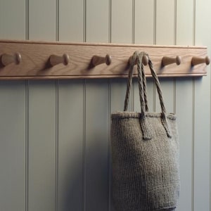 May include: A woven tote bag in a light neutral tone hangs from a wooden coat rack with six pegs. The rack is mounted on a wall with vertical paneling. The bag has rope handles and a textured appearance, suggesting a natural fibre.
