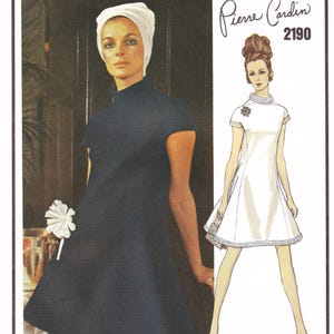 May include: A woman in a dark blue dress and white head covering. The dress has short sleeves and a high neckline. A drawing of a white dress with a high neckline and short sleeves is on the right. The text "Pierre Cardin 2190" is at the top.