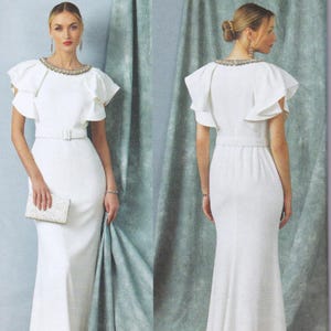 May include: A floor-length white gown featuring short ruffled sleeves, a round neckline adorned with silver embellishments, and a matching white belt at the waist. The dress has a fitted silhouette and is paired with a small, rectangular white clutch.