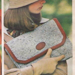 May include: A rectangular clutch bag made of light gray speckled fabric with a dark brown trim along the edges and a central oval brown accent patch on the front flap. The bag features a structured design with a fold-over closure mechanism.