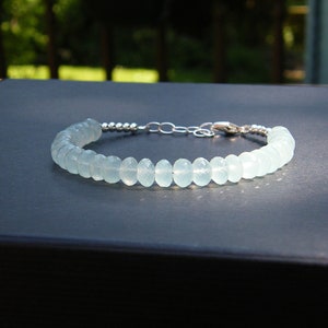 Puede incluir: Una delicada pulsera con cuentas de piedras preciosas facetadas de color azul verdoso pálido. Las cuentas están ensartadas con una cadena y un cierre de plata. La pulsera se muestra sobre una superficie oscura, con un fondo verde borroso.