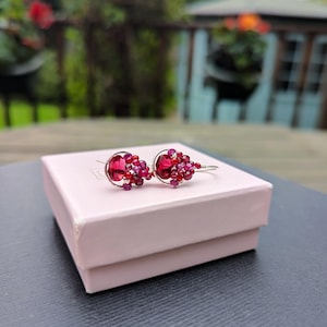 May include: A pair of silver-toned earrings with a cluster of red and pink gemstones. The earrings are resting on a light pink jewelry box. The gemstones are arranged in a floral or berry-like design, with a mix of sizes and shades.
