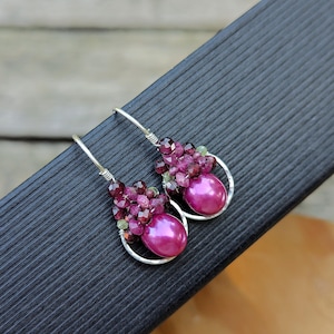 May include: A pair of handcrafted earrings featuring a large, pearlescent, magenta-pink bead at the base, surrounded by clusters of small, faceted, deep red and green beads. The earrings have silver-colored, curved hoops.
