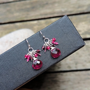 May include: A pair of dangle earrings featuring silver hooks and circular silver rings. Each earring is adorned with a cluster of small, faceted dark pink and red gemstone beads, anchored by a larger, teardrop-shaped faceted red gemstone drop.