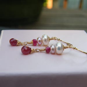 May include: A pair of elegant dangle earrings. Each earring features a teardrop-shaped, faceted ruby at the base, followed by a series of small pink beads, a white pearl, and a larger white pearl. The components are strung on gold-colored wire.
