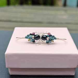 May include: A silver hair barrette with two black, faceted stones at the center, surrounded by clusters of blue, green, and pink beads. The barrette is resting on a light pink box.