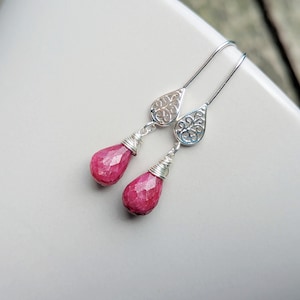 May include: A pair of silver drop earrings featuring faceted, teardrop-shaped pink gemstones. Each earring has a silver wire-wrapped top section connected to a decorative, openwork silver teardrop component that attaches to a long silver ear wire.