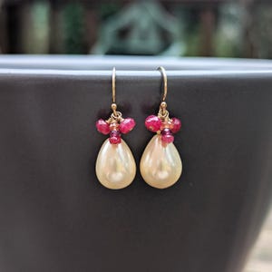 May include: A pair of gold-colored earrings featuring teardrop-shaped pearls. Each earring has a cluster of small, red gemstones at the top, with a gold hook for the ear. The earrings are displayed against a dark background.