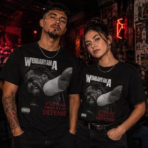 Puede incluir: Dos personas con camisetas negras con un diseño gráfico. Las camisetas presentan el texto "WEMBANYAM A" encima de una imagen de una persona y las palabras "VULGAR DISPLAY OF DEFENSE" debajo. Las camisetas también tienen una advertencia de "CONTENIDO EXPLÍCITO".
