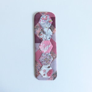 May include: A pink and purple fabric bookmark with a hexagonal pattern. The bookmark is made of different fabrics with floral and geometric designs.
