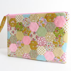 Puede incluir: Bolsa acolchada con diseño patchwork. Presenta una variedad de telas florales y estampadas en tonos rosa, amarillo, verde y azul, dispuestas en un patrón hexagonal. Tiene una cremallera rosa y un tirador de tela a juego.