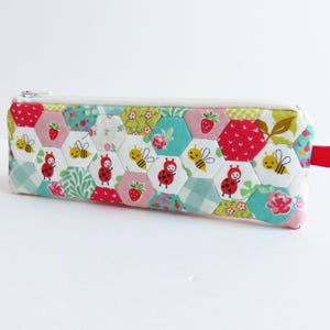 May include: A white zippered pencil case with a patchwork design featuring hexagons in shades of pink, red, green, blue, and white. The hexagons are decorated with small red ladybugs and yellow bees.