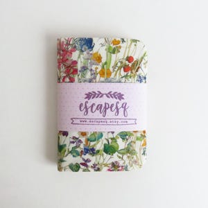 May include: A floral patterned notebook with a light purple band featuring the text "escapesq" and a website address. The notebook's cover displays a variety of colorful flowers and greenery against a white background.