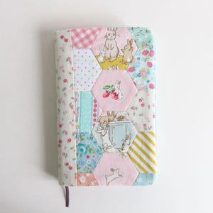 May include: A quilted book cover with a patchwork design featuring bunnies, strawberries, and floral patterns. The cover has a pink, blue, and white color scheme with a purple ribbon bookmark. The cover is rectangular and has a soft, handmade appearance.