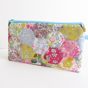Puede incluir: Una bolsa de cremallera de tela patchwork con una cremallera azul y un patrón hexagonal. La bolsa está hecha de varias telas florales en tonos de rosa, amarillo, verde y azul.